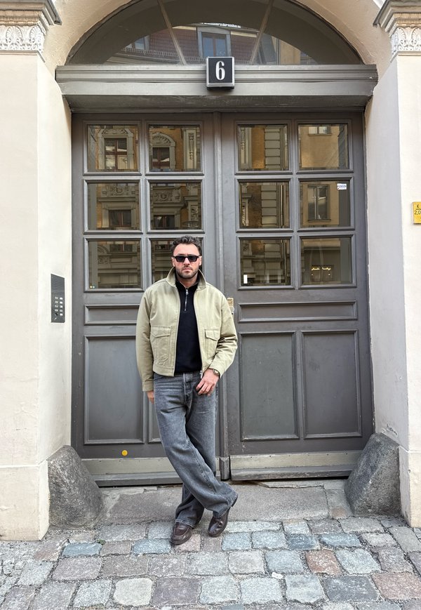 Mann mit Sonnenbrille, beiger Jacke und Jeans steht mit überkreuzten Beinen vor einer großen grauen Tür mit der Nummer 6 auf einer Kopfsteinpflasterstraße.