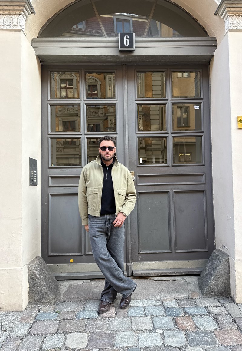 Homme portant des lunettes de soleil, une veste beige et un jean, debout les jambes croisées devant une grande porte grise numérotée 6, sur une rue pavée.