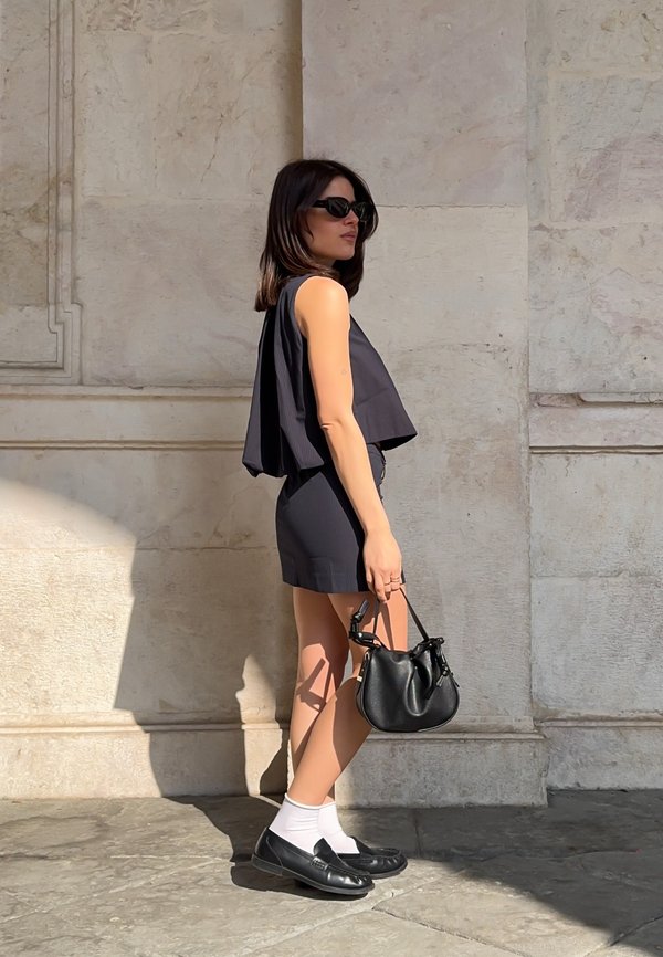 Mujer con top sin mangas negro y pantalones cortos, gafas de sol negras, calcetines blancos, zapatos negros, sosteniendo un bolso negro, de pie contra una pared de piedra beige.