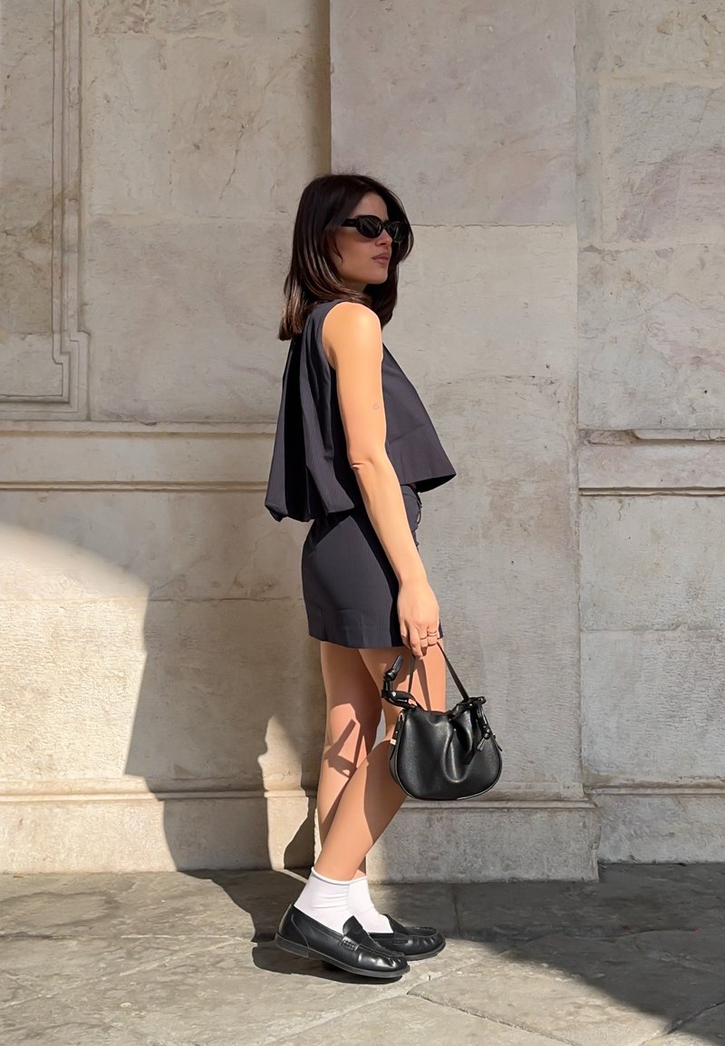 Donna con top nero senza maniche e pantaloncini, occhiali da sole neri, calzini bianchi, scarpe nere, che tiene in mano una borsa nera, in piedi contro un muro di pietra beige.