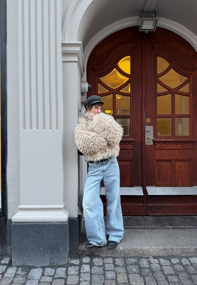 Chaqueta de piel sintética de color crema esponjosa, combinada con jeans sueltos de color azul claro y botas negras, apoyada contra una columna de piedra cerca de unas puertas de madera.