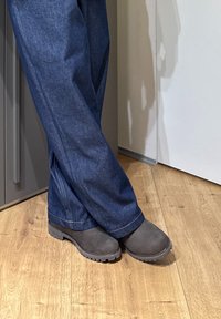Personne portant un jean bleu foncé et des bottes gris foncé, debout jambes croisées sur un sol en bois près d'une porte blanche.