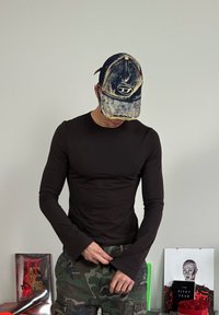 Svart långärmad tröja med figurnära design och vida ärmslut, tillsammans med kamouflage cargo-byxor och en sliten denimkeps med en logotyp.