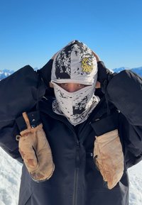 Gorro e máscara com padrão preto e branco, combinados com uma jaqueta preta. Luvas castanhas mostram sinais de uso. Fundo nevado, céu azul claro.