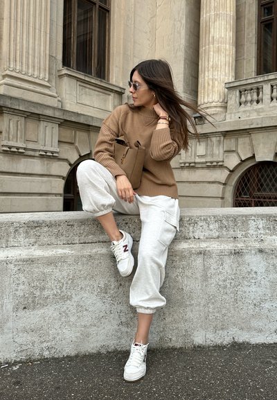 Suéter marrón con canalé, pantalones de chándal grises y zapatillas blancas con detalles rojos. Sosteniendo una bolsa marrón, de pie sobre un muro de piedra junto a un edificio.