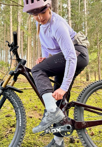 Bicicleta de montaña con un marco color marrón oscuro, horquilla de suspensión dorada y neumáticos anchos. Ciclista con una camiseta de manga larga de color púrpura claro y pantalones negros.