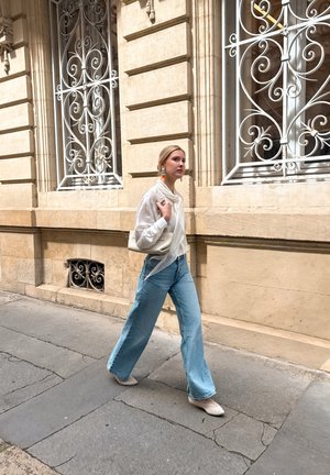 Mujer caminando por la calle con una blusa blanca, vaqueros anchos azul claro, zapatos blancos y llevando un bolso blanco cerca de un edificio de piedra beige con ventanas ornamentadas.