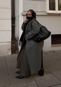 Grijze wollen jas, zwarte sjaal en oorwarmers. Zwarte handtas met studs. Zwarte puntige schoenen. Staand op het trottoir tegen een muur.