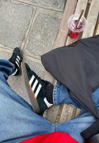 Groene suède sneakers met witte strepen, zwarte accenten en een bruine zool. Blauwe denim jeans en een zwart item naast een rode shirt.