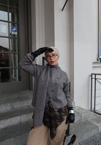 Femme portant un foulard beige, un pull gris, un pantalon avec détails en dentelle noire et des gants noirs, debout sur des marches en pierre près de l'entrée d'un bâtiment.