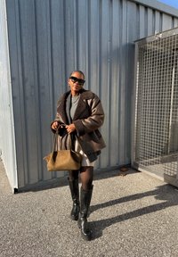Femme portant des lunettes de soleil, veste en shearling marron, bottes noires, tenant un sac à main beige, debout sur du béton près d'un mur industriel en métal et d'une porte.