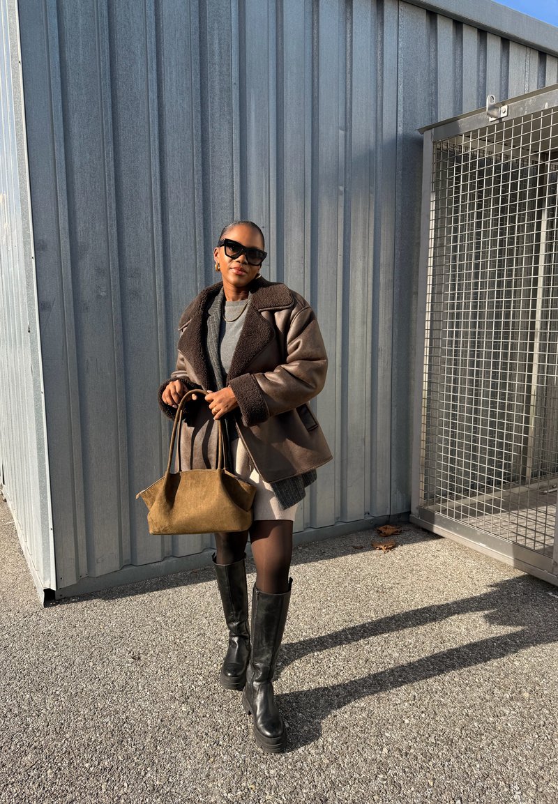 Kvinde iført solbriller, brun shearlingjakke, sorte støvler, holder en tanfarvet håndtaske, stående på beton nær metal industrielt væg og port.