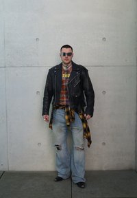 Svart skinnjakke over en rutete skjorte, slitte lyseblå jeans, gul rutete skjorte bundet rundt hoften, sølvsmykker og solbriller.