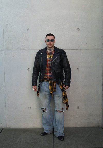 Chaqueta biker de cuero negro sobre una camisa de cuadros, vaqueros azul claro desgastados, camisa de cuadros amarilla atada a la cintura, accesorios plateados y gafas de sol.