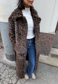 Manteau en fausse fourrure imprimé léopard avec col châle, porté sur un pull blanc côtelé et un jean flare bleu, associé à des mocassins beiges.