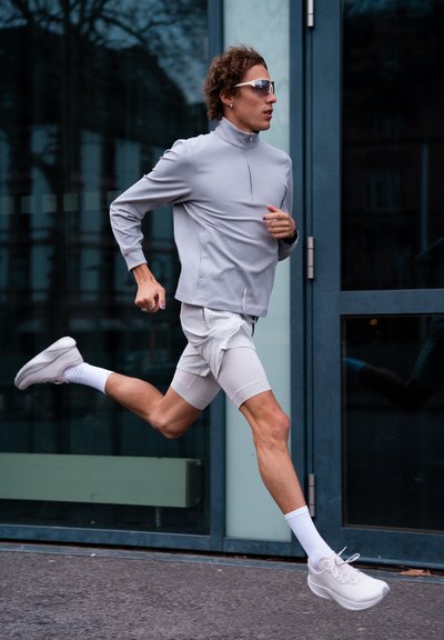 Joven con ropa deportiva gris y gafas de sol corriendo junto a puertas de cristal en una acera urbana durante el día.