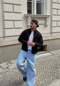 Fekete gombos ing fehér póló felett, világoskék, bőszárú farmerrel és fekete cipővel párosítva. Városi környezet, macskaköves járda.