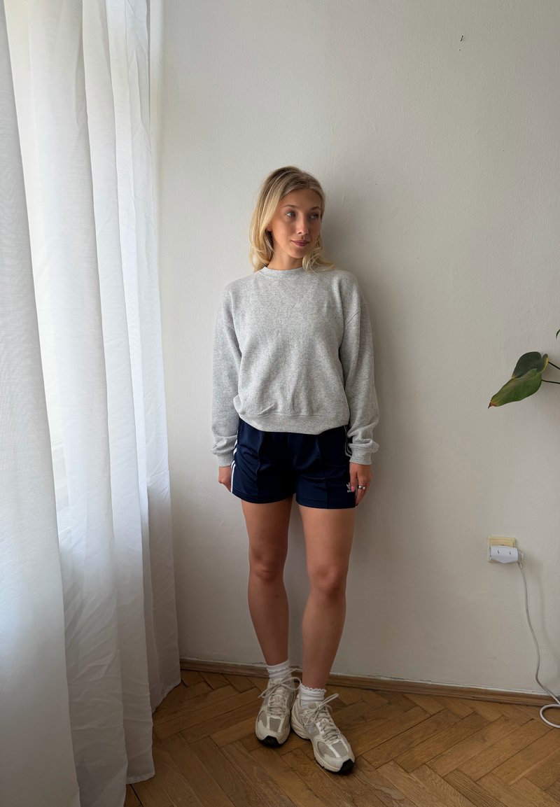 Femme debout à l'intérieur portant un sweat gris, un short de sport bleu marine, des chaussettes blanches et des baskets, à côté d'un rideau et d'un mur blancs.