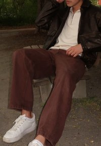 Brune kordfløjspants, hvide snøresneakers, tekstureret hvid top med kvart lynlås og en mørk læderjakke. Siddende på en træbænk.
