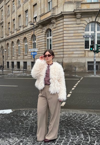 Mujer ajustándose las gafas de sol, vestida con chaqueta blanca de piel sintética, camisa roja a rayas y pantalones anchos beige, de pie en una esquina de una calle adoquinada nevada.