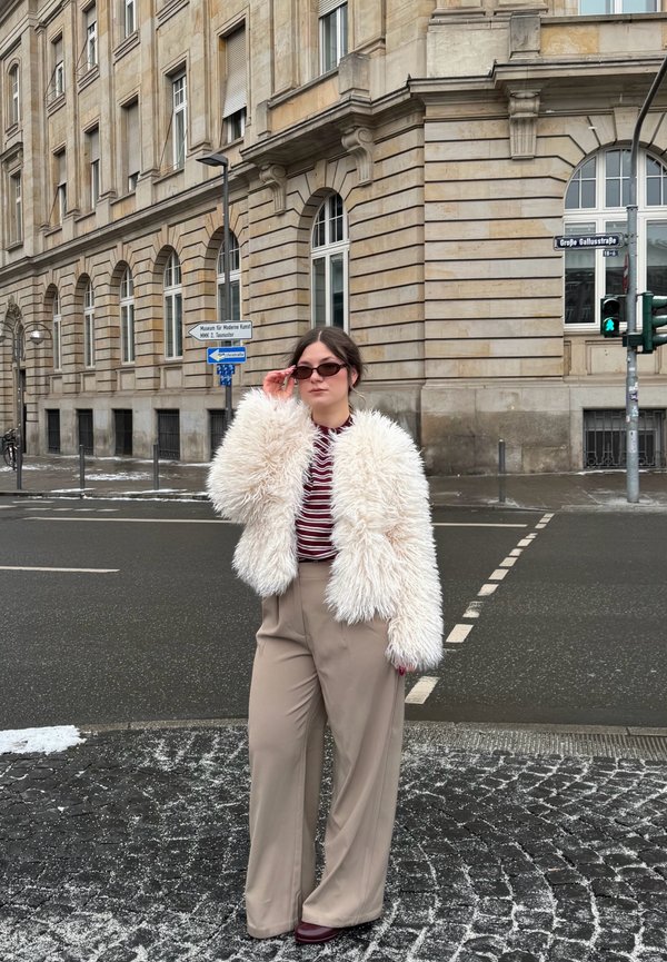Donna che si sistema gli occhiali da sole indossando una giacca in pelliccia ecologica bianca, una camicia a righe rossa e pantaloni larghi beige, in piedi all'angolo di una strada di ciottoli innevata.