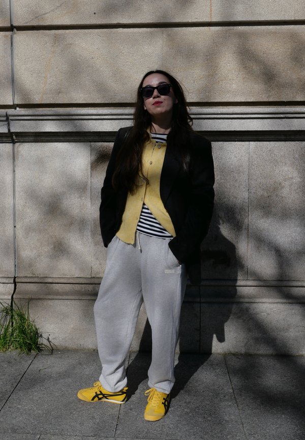 Kvinde iført solbriller, sort blazer, gul cardigan, stribet skjorte, grå joggingbukser og gule sneakers, stående foran en stenmur i dagslys.