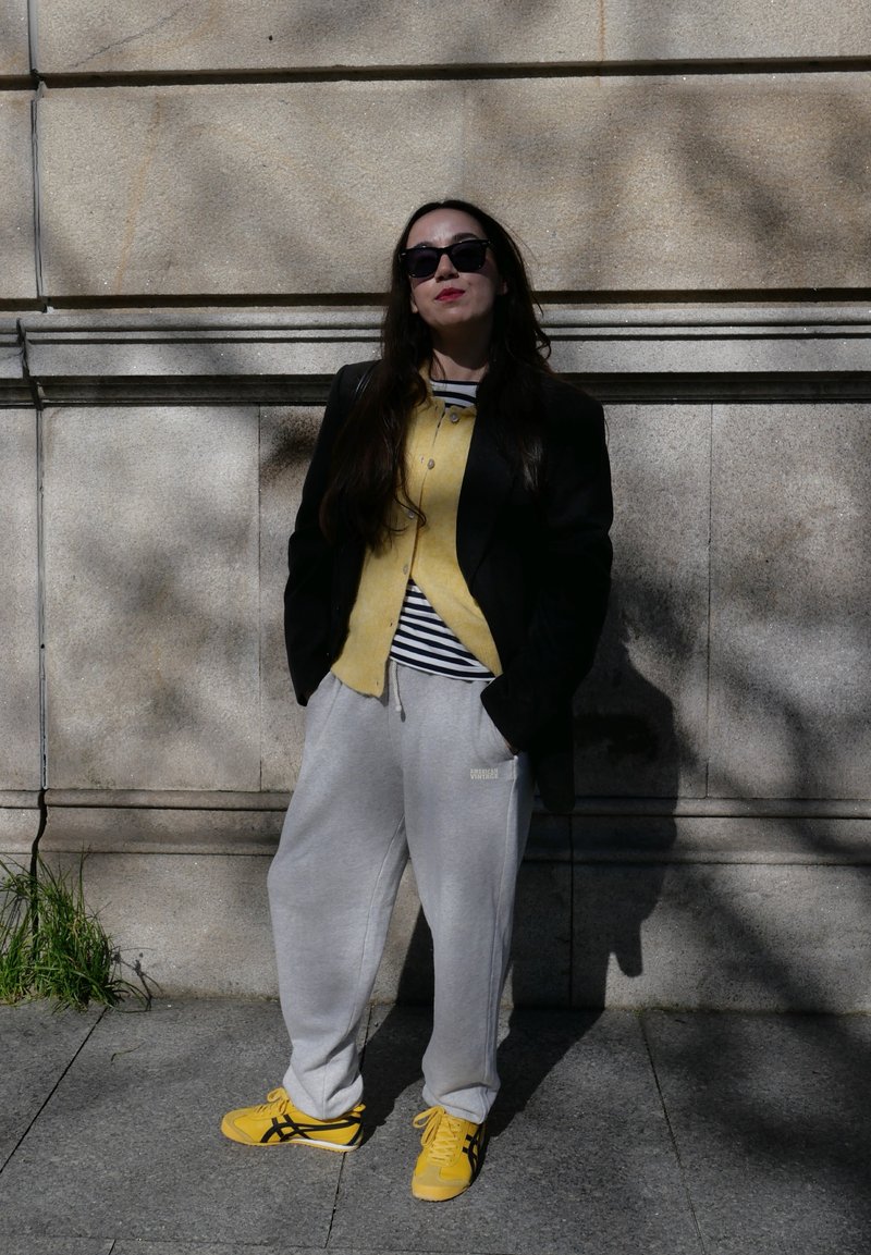 Femeie purtând ochelari de soare, sacou negru, cardigan galben, cămașă în dungi, pantaloni sport gri și adidași galbeni, stând în fața unui zid de piatră, în lumina zilei.