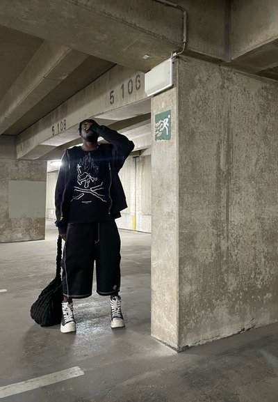 Sudadera negra con diseño gráfico, pantalones cortos negros, zapatillas altas blancas y una bolsa negra texturizada, en un garaje de estacionamiento de concreto.