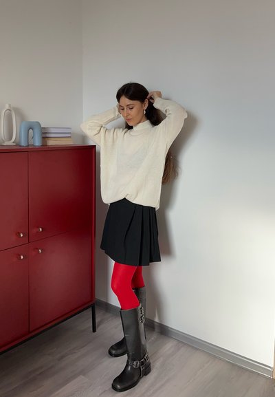 Suéter de punto color crema con cuello alto, falda negra plisada, medias rojas, botas negras hasta la rodilla con hebillas. Armario rojo y decoración minimalista de fondo.