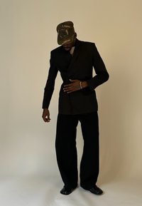 Mann in einem schwarzen Anzug und weitem Hosen, mit einer camouflagemuster Mütze und schwarzen gespaltenen Schuhen, vor einem schlichten beigen Hintergrund stehend.