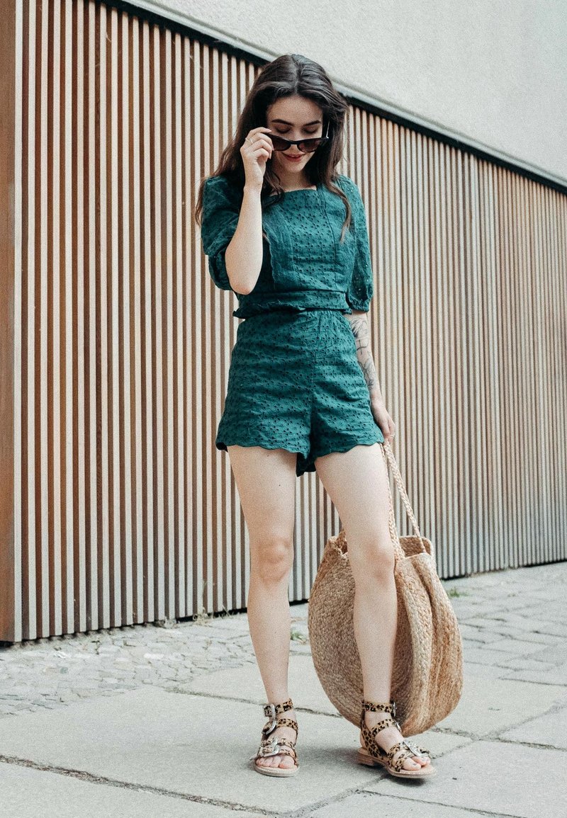 Zöld textúrájú ruha hullámos szélekkel, barna szőtt táskával és állatmintás szandálokkal, fa lamellás háttér előtt.