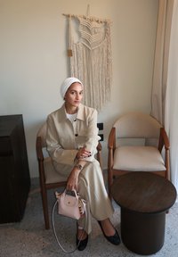 Costum bej cu dungi, cu o croială lejeră, completat de un batic alb. Stând pe un scaun din lemn lângă o masă rotundă întunecată, cu o geantă mică roz.