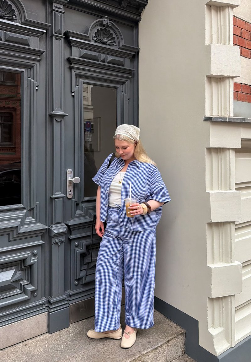 Femme en tenue à carreaux bleu et blanc tenant un café glacé, debout à côté d'une porte ornée gris foncé dans une rue de la ville.