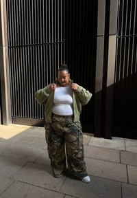 Jachetă bomber verde peste un top alb ajustat, asociată cu pantaloni cargo camuflați. Setările au fâșii verticale negre și un podea din piatră.
