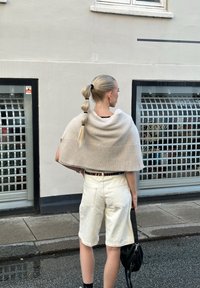 Ανοιχτό μπεζ πλεκτό πόντσο με ριγωτή στρίφωμα, φορεμένο πάνω από ένα μαύρο τοπ, συνδυασμένο με λευκά τζιν σορτς και μαύρες κάλτσες μέχρι τον αστράγαλο, κρατώντας μία μαύρη τσάντα.