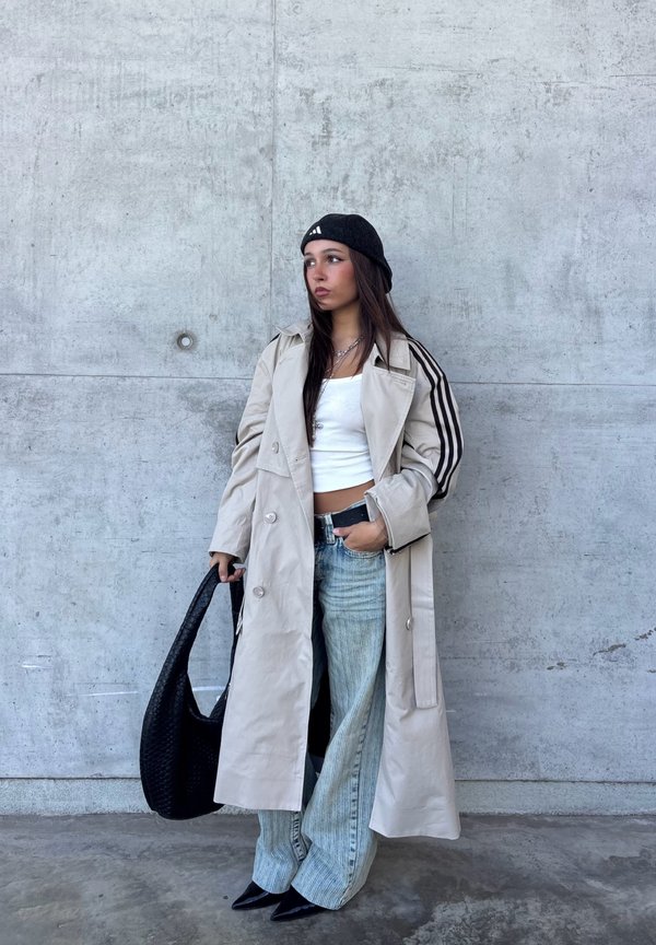 Joven mujer con gabardina beige, top corto blanco, vaqueros azul claro, boina negra y zapatos negros, de pie contra una pared de hormigón sosteniendo un bolso negro.