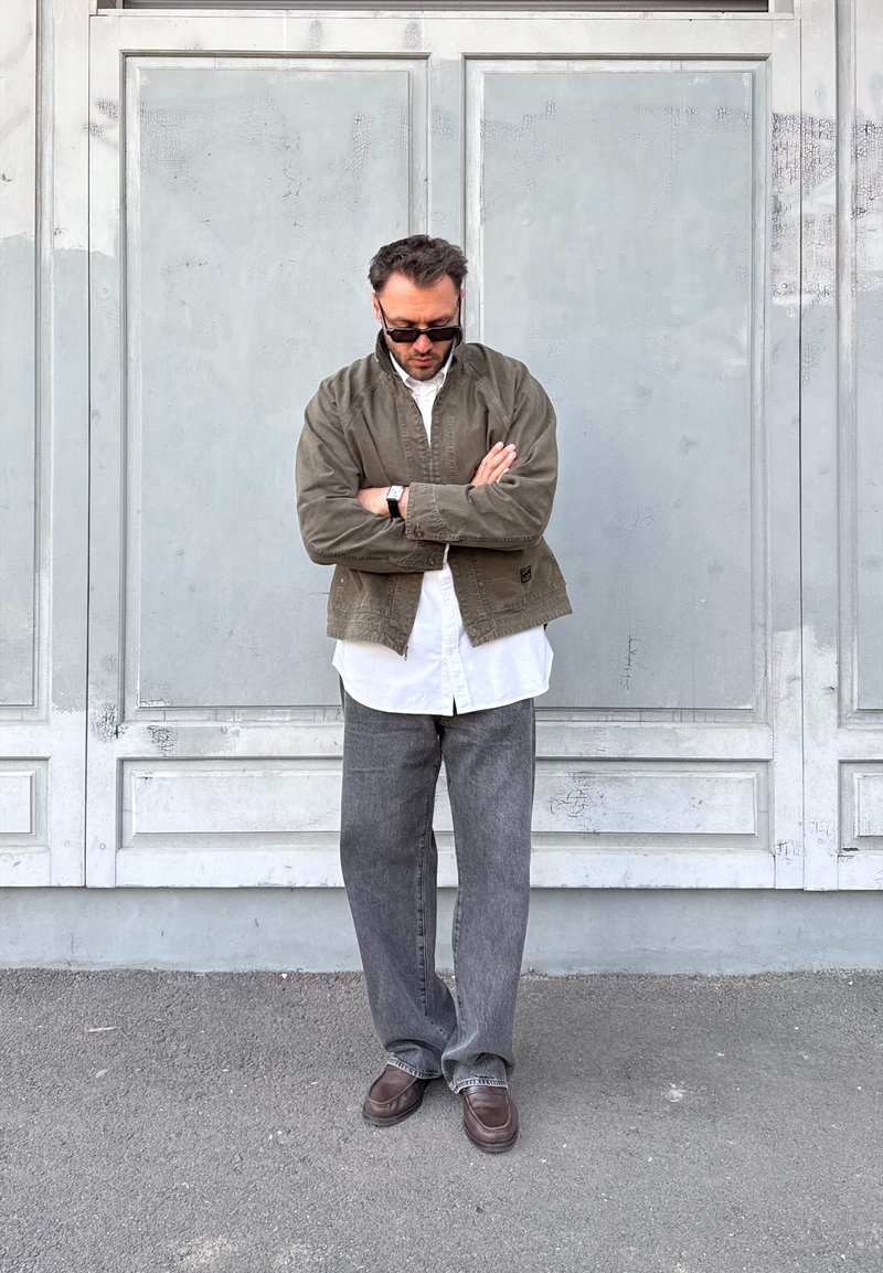 Homme portant des lunettes de soleil, une veste verte, une chemise blanche et un pantalon gris, debout les bras croisés contre un mur à panneaux gris.