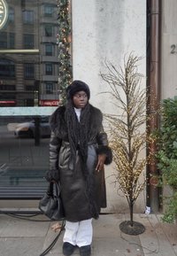 Lang zwart leren jas met bontaccenten, witte coltrui, zwarte handschoenen en een bijpassende bontmuts. Donkere tas en witte broek maken de outfit compleet.