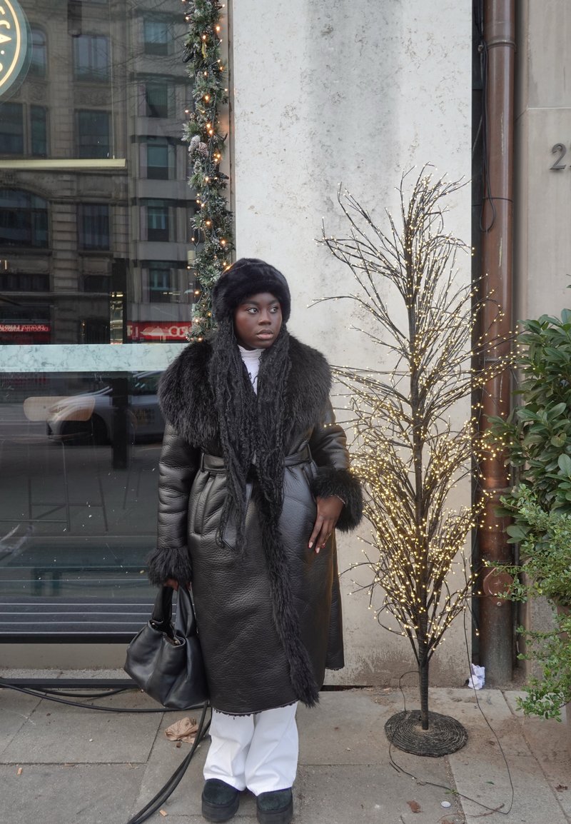 Long manteau en cuir noir avec accents en fourrure, col roulé blanc, gants noirs et un chapeau en fourrure assorti. Sac foncé et pantalon blanc complètent la tenue.