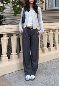 Ανοιχτό γκρι πλεκτό κασκόλ πάνω από λευκό μπλουζάκι, συνδυασμένο με χαλαρές σκούρες γκρι παντελόνες και λευκά αθλητικά παπούτσια με γκρι λεπτομέρειες.