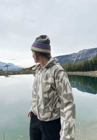 Gai pelēka jaka ar ģeometrisku rakstu, kopā ar krāsainu cepuri. Fona ir ainavisks ezers un kalni.