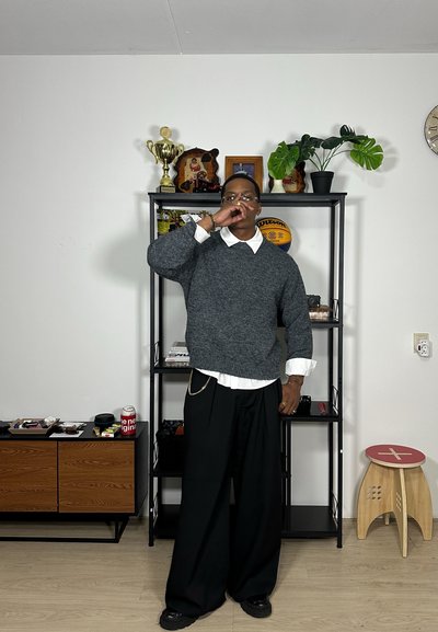 Suéter gris de punto sobre una camisa blanca con cuello, combinado con pantalones anchos negros. Los accesorios incluyen una cadena plateada y gafas.