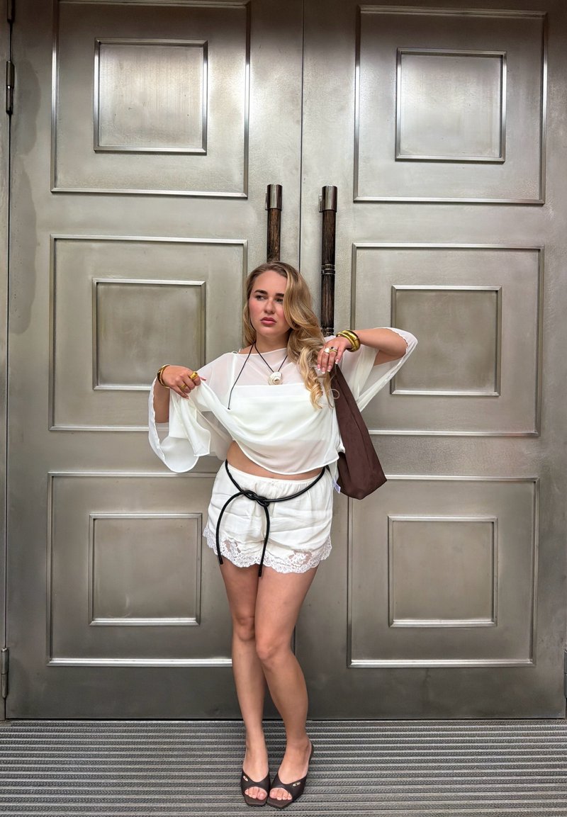 Femme blonde portant un haut transparent blanc et un short avec une ceinture noire, debout les bras levés devant de grandes portes doubles métalliques.