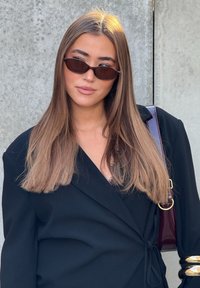 Melns wrap stila žakete ar jostu viduklī, saskaņots ar mazām brūnām saulesbrillēm. Gara, taisna brūna matu krāsa un spīdīga violeta soma.