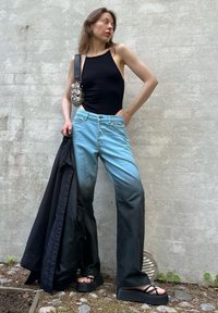 Top de alças justo preto, jeans de denim azul claro a escuro, sandálias de plataforma pretas e uma pequena bolsa estampada. Em pé contra uma parede cinza.