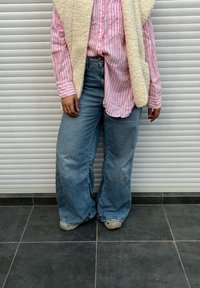 Camisa de botões às riscas em rosa claro e branco combinada com um colete de pelo falso creme. Jeans azuis de ganga de corte largo e solto, com calças de perna larga, e ténis cinzentos.