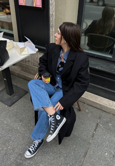 Blazer negro de gran tamaño, camisa de mezclilla, vaqueros azul claro y zapatillas altas negras de Converse. Sosteniendo una pequeña taza de café amarilla. Entorno al aire libre.