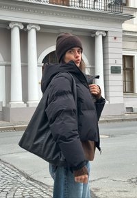 Doudoune noire à capuche, coupe courte, associée à un pull en maille marron. En arrière-plan, des colonnes en pierre et la façade d'un bâtiment.