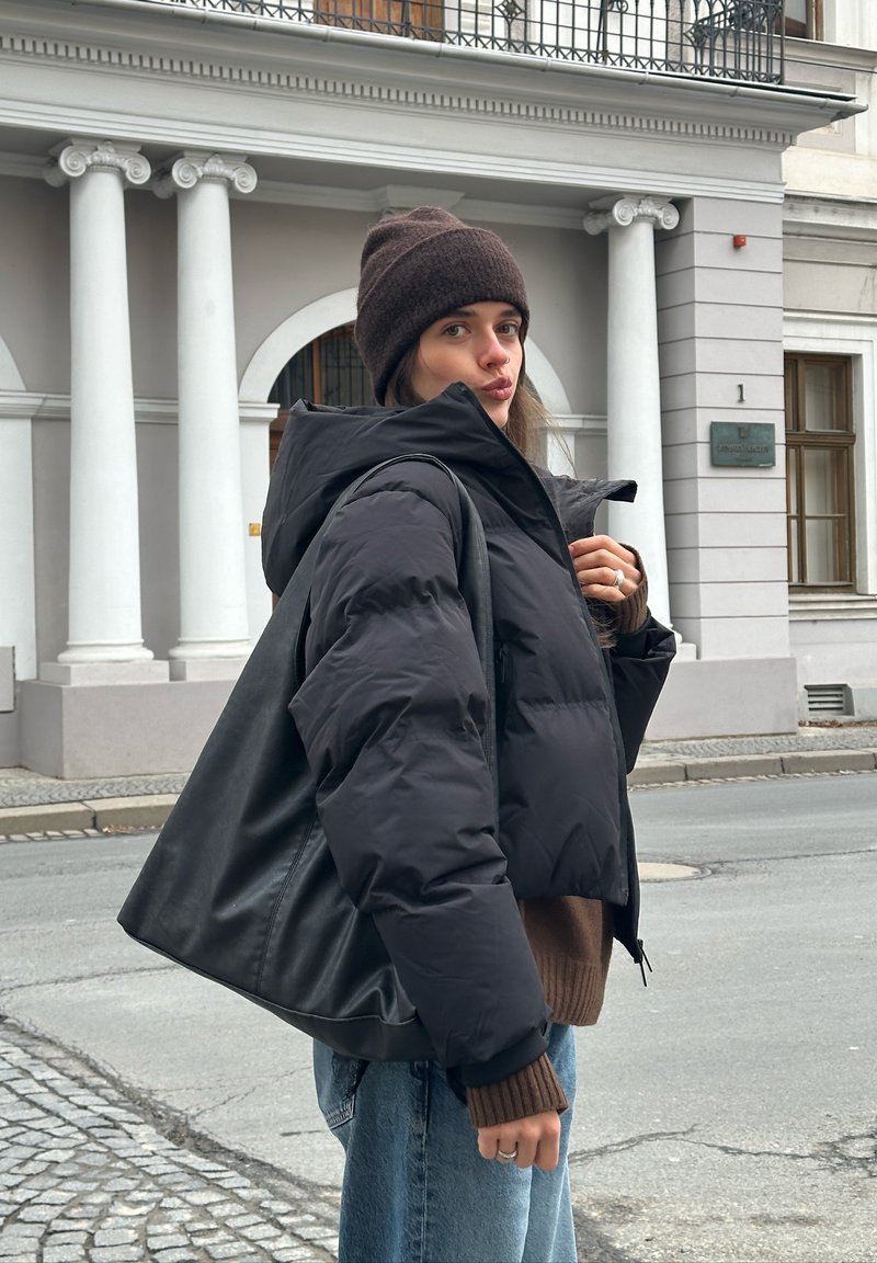 Juoda pūkinė striukė su kapišonu, trumpintas dizainas, derinama su rudu megztiniu. Fone matyti akmeninės kolonos ir pastato fasadas.