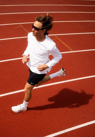 Corredor con una camiseta de manga larga blanca y pantalones cortos negros, usando gafas de sol, corriendo en una pista roja con marcas de carril blancas. Las zapatillas deportivas tienen acentos rojos.